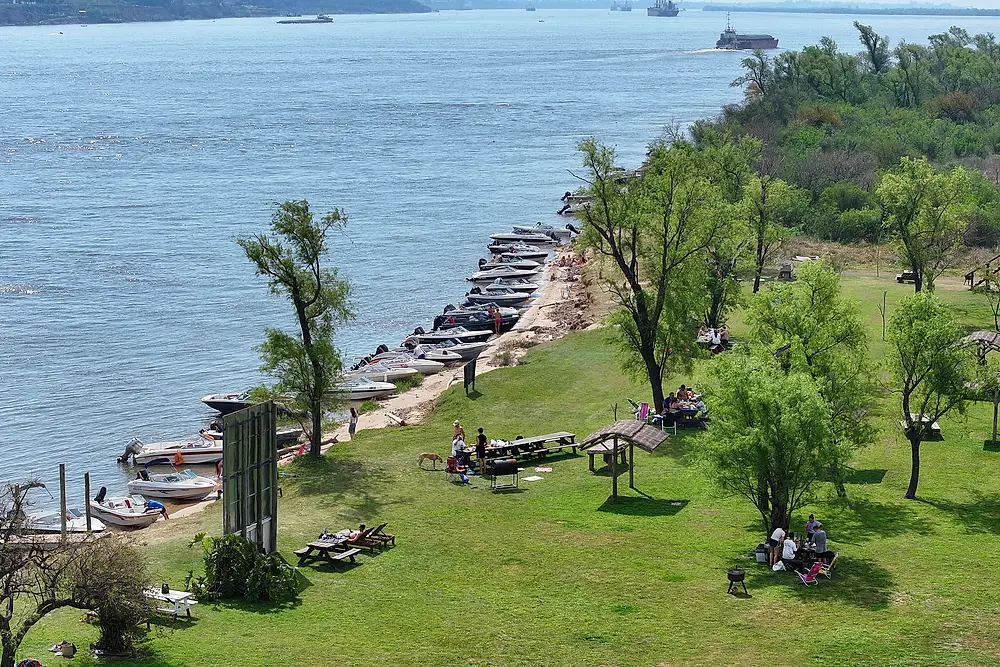 Parque con árboles y mesas junto a la costa, con lanchas amarradas sobre el río